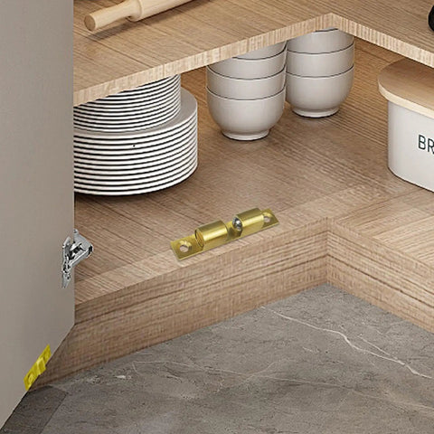 Adjustable Roller Ball Door Catch For Cabinets, Closets, And Wardrobes on a wooden shelf next to plates, brass finish