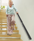 Wall Mounted Stair Handrail Crafted From Premium Stainless Steel Featuring A Sleek Black Square Profile Built Like A Tank For Indoor Spaces Loaded With Rock Solid Grip And Holds Down Weighty Loads Up To Two Hundred Pounds Safely, shown in use by elderly woman on staircase.
