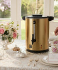 Tabletop setup with a gold 6L Coffee Urn Hot Beverage Dispenser Electric Coffee Percolator With Double Wall Stainless Steel 38 Cup Capacity Commercial Grade Large Coffee Maker For Party Home Catering Gold Color next to floral teacups and glasses.