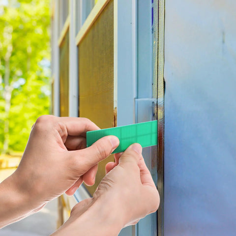 Plastic flat shim, green, installed between blue window frame and wall for leveling.