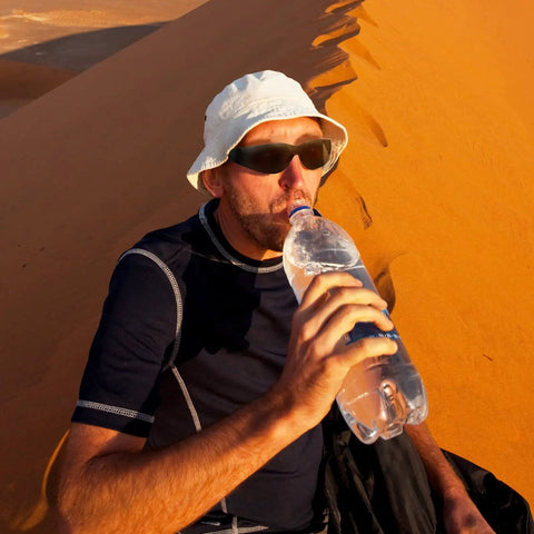 Person wearing sunglasses and hat drinking water in sunny desert environment
