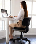 Woman sitting at desk using laptop with Upgraded Memory Foam Coccyx Cushion For Office Chair And Driving Tailbone Pain Relief.