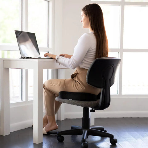 Woman sitting at desk using laptop with Upgraded Memory Foam Coccyx Cushion For Office Chair And Driving Tailbone Pain Relief.