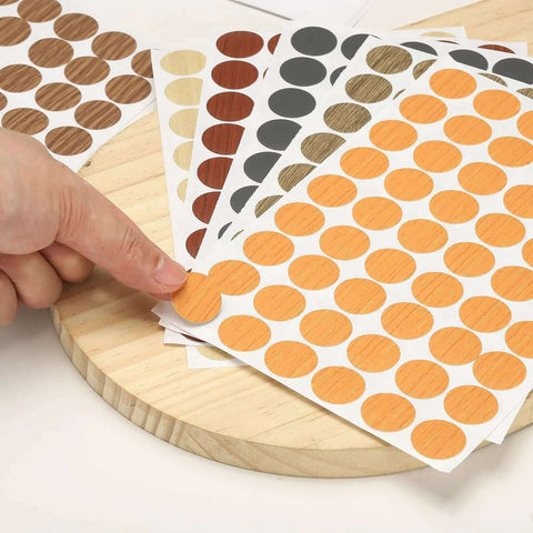 Heavy Duty Self Adhesive Screw Hole Caps 540 Pack Slick Yellow Wood Grain Finish on sheets with finger pointing to cap.