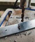 Close-up of a cobalt drill bit drilling into a metal strip on a workbench with metal shavings.