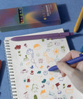 Person drawing with Colorful Gel Pen Set, 6 Pack Fine Point 0.5mm Colored Ink Pens For Journaling, on notebook with doodles.