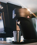 Game Changing Single Dose Coffee Grinder With 64 Millimeter Flat Burrs Stepless Adjustment Heavy Duty Zero Retention Build For Killer Espresso At Home Quiet Compact Design In Crisp White Finish placed beside an espresso machine.