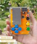 Hand holding the Hands On Solder Kit With Five Retro Game Classics For Electronics Practice And Learning Handheld Size In Clear Acrylic Case Easy To Build And Play Great Gift For Family And Friends, showing buttons and digital display in a garden.