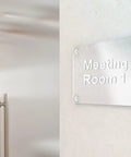 Clear acrylic meeting room sign with screws on wall showcasing "Meeting Room 1" text.