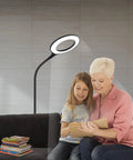 Gooseneck LED Floor Lamp For Office Reading And Bedrooms Non Glare Eye Safe Lighting 600 Lumens From 3 Watt LED Energy Saver Lit Brightness Ideal For Lash Extensions Makeup And Nail Spa Black, illuminating a reading area with two people sharing a book on a sofa.