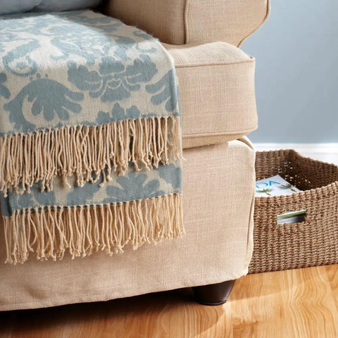 Beige armchair with decorative blue and beige fringed throw blanket, positioned on hardwood floor near woven basket.