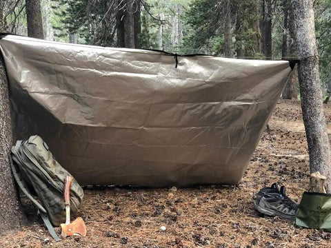 Rock Solid Insulated Tarp And Thermal Survival Blanket, Shuts Out Hard Weather While Locking In Ninety Percent Body Heat For Campouts And Bugouts At Sixty By Eighty Two Inches With Reinforced Grommets set up in a forest with a backpack and axe nearby.