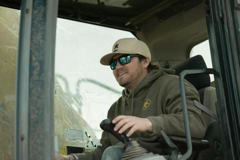 Kickass Z87 Certified Wayfarer Safety Sunglasses Featuring Hella Effective Antifog Smoke Lenses And Totally Shatterproof Matte Black Frames Built Tough For Male Workers, worn by a worker in a vehicle.