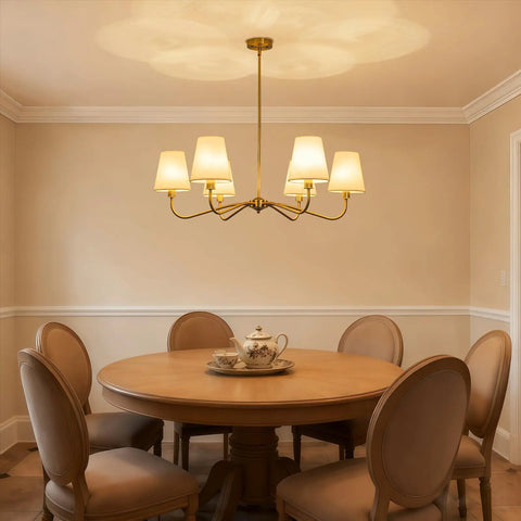 Smooth Operator 6 Light Brass Gold Chandelier With White Fabric Shades above round dining table in cozy setting.