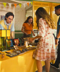People shopping at a stall with various items on a yellow table in a market setting.