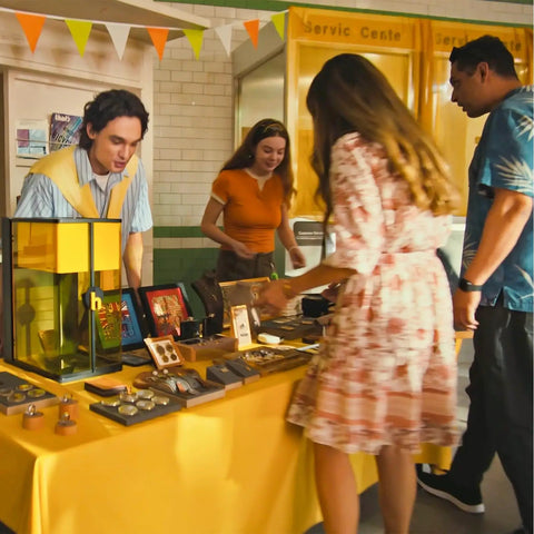 People shopping at a stall with various items on a yellow table in a market setting.