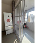 Custom Sized Insulated Sliding Accordion Room Divider With Smooth Folding Panels And Zero Sweat Setup For Interior Patios Or Laundry Rooms Featuring Heavy Duty Waterproof Aluminum Frame In Bright White next to a refrigerator in a bright room.