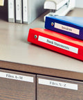 Slick Portable Monochrome Label Maker Featuring Qwerty Keyboard And Crisp Display Designed For Home Office Organization Includes Four Nifty Tape Rolls For Dead Simple Everyday Printing Tasks seen labeling red and blue binders on a desk.