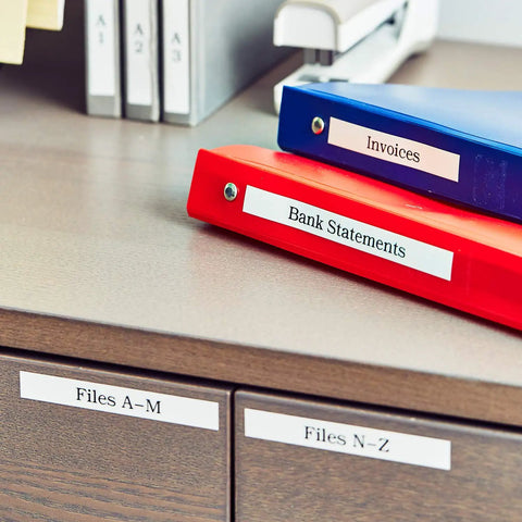 Slick Portable Monochrome Label Maker Featuring Qwerty Keyboard And Crisp Display Designed For Home Office Organization Includes Four Nifty Tape Rolls For Dead Simple Everyday Printing Tasks seen labeling red and blue binders on a desk.