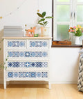 White dresser with decorative blue tile patterns in bright room, beside a window with plants and flowers.