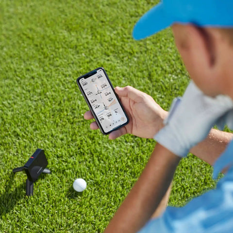 User checking swing metrics on phone next to Portable Golf Launch Monitor And Simulator Bundle With 10 Hour Battery Life on grass