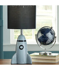 Kids Rocket Table Lamp With Gray Ceramic Base And Fabric Drum Shade beside a globe on a desk