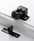 Close-up of twin roller bronze door catch from Pack Of 5 Heavy Duty Plated Door Catches With Screws on white cabinet edge.