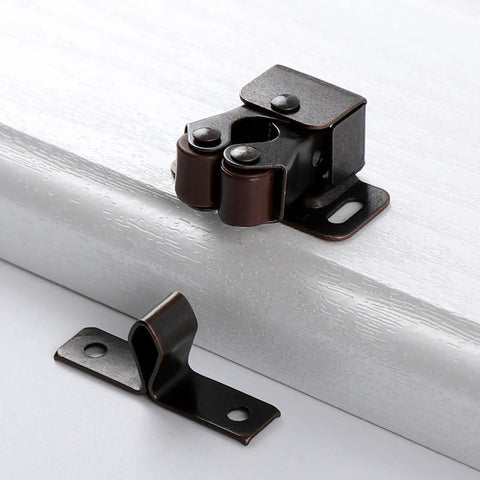 Close-up of twin roller bronze door catch from Pack Of 5 Heavy Duty Plated Door Catches With Screws on white cabinet edge.