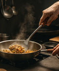 Side view of a stainless steel wok spatula with wooden handle lifting stir-fry in a metal wok over a gas stove.