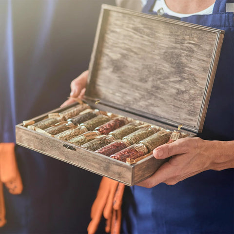 Person holding a wooden box with glass vials organized in rows, showcasing assorted spices and herbs. Box is open with visible hinges.