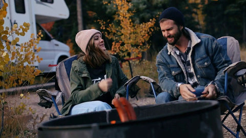 Campers sitting by fire with RV in background, enjoying evening outdoors.