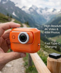 Hand holding 4K Magnetic Action Camera With 48MP Sensor Dual Battery, orange, in mountain setting with charging feature details visible.