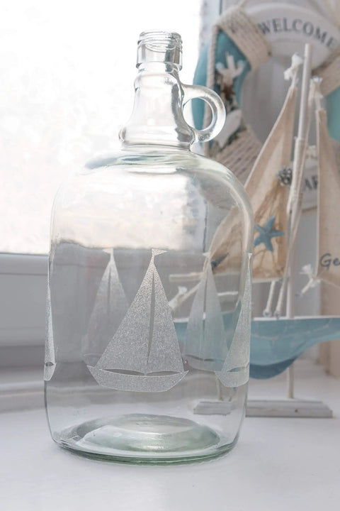Clear glass jug with engraved sailboat design, nautical-themed background with decorative boat and welcome sign.