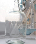 Clear glass bottle with etched sailboat design, nautical decor in background with model sailboat and welcome sign.
