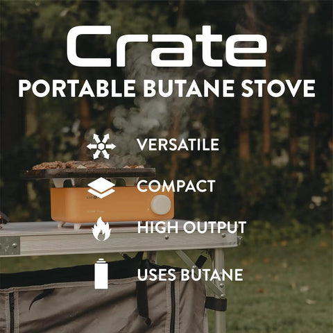 Crate Series butane camp stove on a table outdoors, showing its compact size and orange color. Text highlights features: versatile, compact, high output, uses butane.