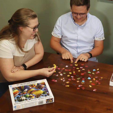 Two people assembling the Hot Air Balloon Festival 1000 Piece Jigsaw Puzzle With Easy Grip Pieces on a table.