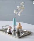 Three decorative ceramic vases in pastel pink, turquoise, and beige with metallic silver leaf bases on a silver tray.