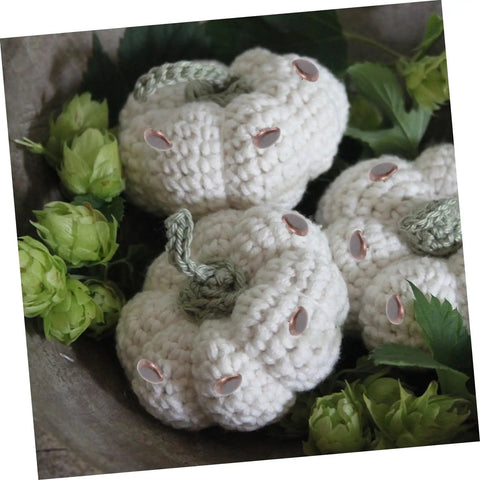Crocheted white pumpkins with green stems and embellishments surrounded by green foliage, displayed in a decorative setting.