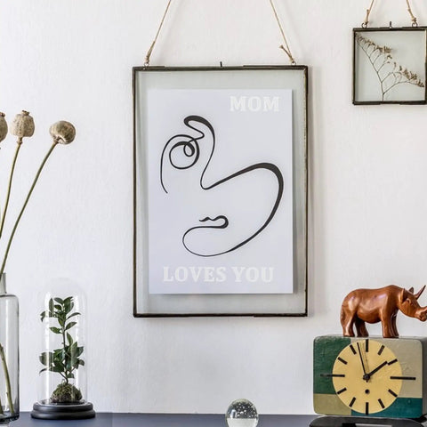 Minimalist wall art "Mom Loves You" with abstract face print in frame, on a shelf with decor including a rhino statue and clock.