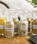Glass jars filled with various spices, including chai, spicy salt, and curry, labeled on a wooden table with a notebook nearby.