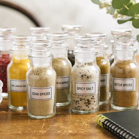 Glass jars filled with various spices, including chai, spicy salt, and curry, labeled on a wooden table with a notebook nearby.