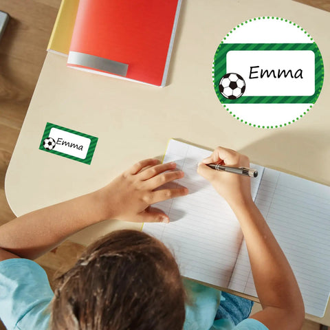 Child writing at desk with 400 Soccer Name Tag Stickers Set In Green And Black With 4 Designs. Sticker labeled 'Emma'.
