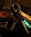 Embrite Glow Fob with Green Embrite Pellet glowing on keychain with watch and pocket knife in a dark setting