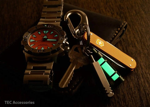 Embrite Glow Fob with Green Embrite Pellet glowing on keychain with watch and pocket knife in a dark setting