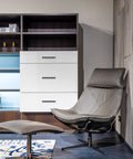 Modern living room with leather recliner chair and footstool, white cabinet with three drawers, shelf unit in sleek design.
