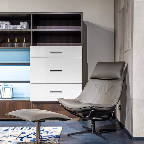 Modern living room with leather recliner chair and footstool, white cabinet with three drawers, shelf unit in sleek design.