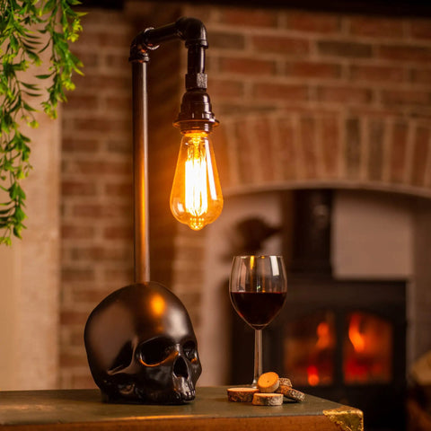 Black Industrial Skull Lamp With Skeleton Decor For Gothic Bedroom Dope Handcrafted Cast Iron And Resin Skull Table Lamp Vintage Industrial Vibe UL Certified 18.5 Inches Tall 1 to 40W Bulb Not Included next to a wine glass on a rustic table