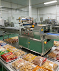 Sleek Compact Horizontal Flow Wrapper Shrink Wrap Sealer Featuring Dead Simple Setup And No Sweat Operation Packing Fifty To Eighty Units Per Minute For Deli Shops And Fresh Markets on a countertop with packaged food.