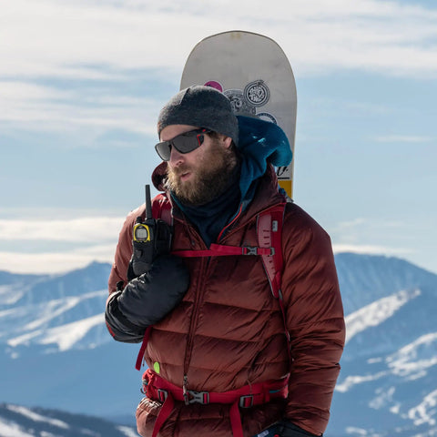 Man using Push To Talk Handheld And Wearable Speaker Microphone Set For GMRS Radios, with snowboard, in snowy mountain setting