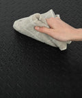Close-up of black diamond plate design on Heavy Duty Anti Fatigue Floor Mat Built For Long Shifts 3 Ft By 7 Ft Thick Fifteen Sixteenths Inch Ergonomic Standing Mat For Commercial Kitchens And Workshop Floors being cleaned with a cloth.